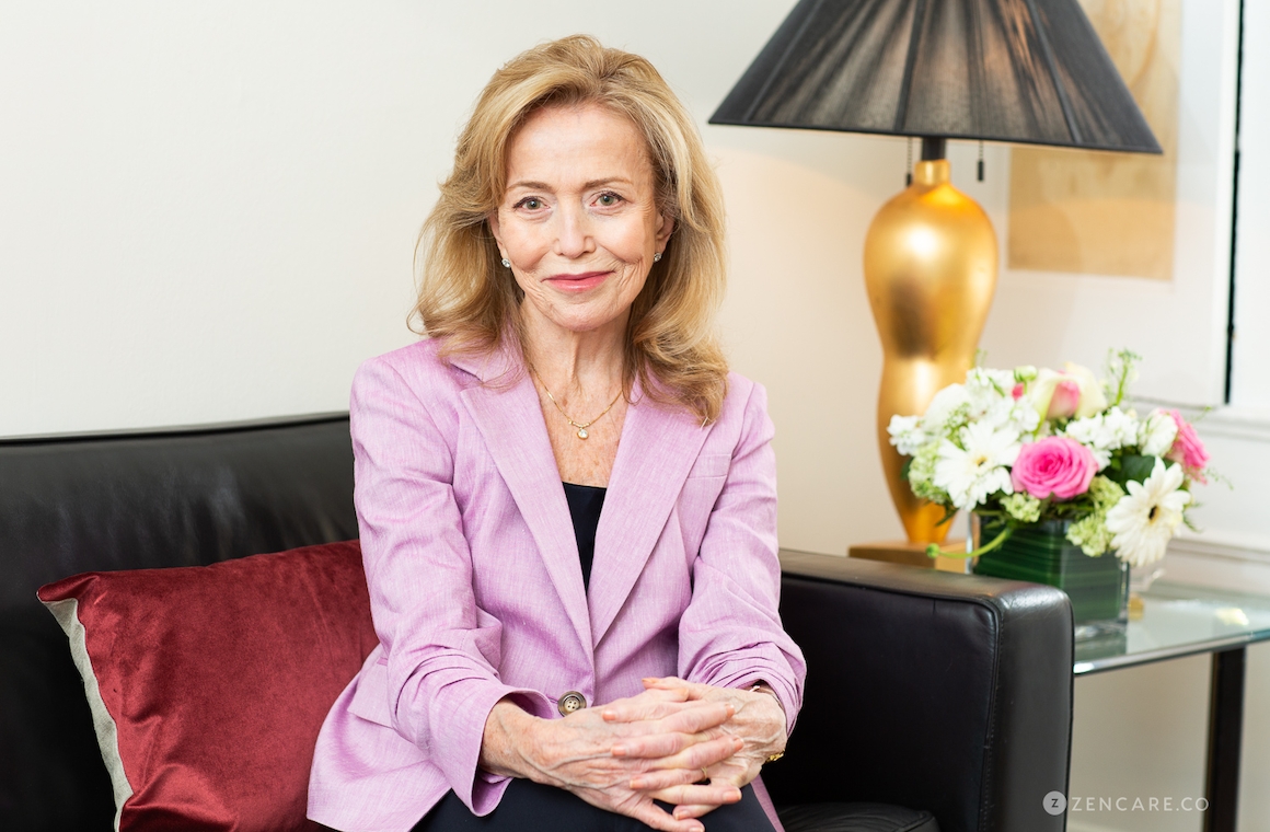 Dr. Deborah Grody, a licensed clinical psychologist in New York City, seated in her office, wearing a light purple blazer, offering a warm and welcoming expression with a backdrop of a lamp and floral arrangement.